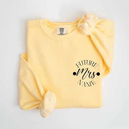 May include: A light yellow sweatshirt with the text "FUTURE Mrs NAME" in black script. The sweatshirt is folded with the sleeves tied in a knot. The Comfort Colors brand tag is visible.