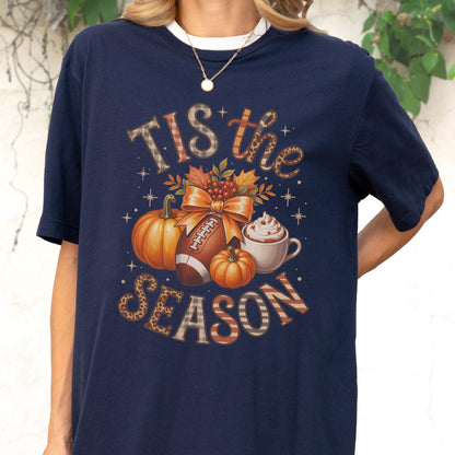May include: Navy blue t-shirt with a fall-themed graphic. The design includes pumpkins, a football, a bow, and a cup of whipped cream. The text "Tis the Season" is displayed in a mix of fonts and patterns.
