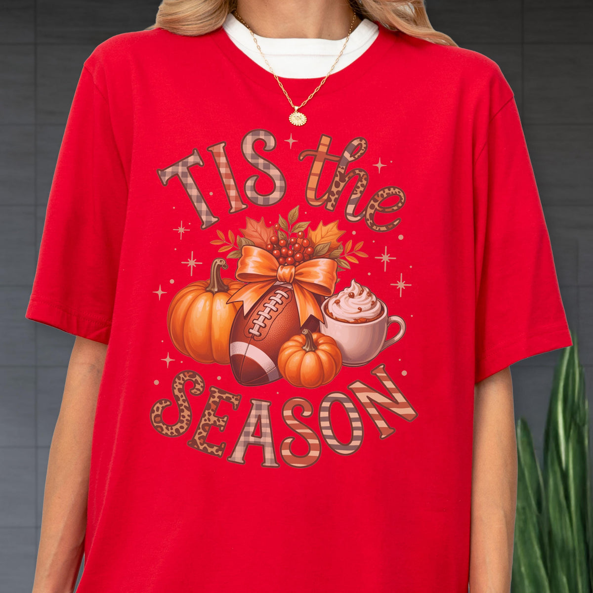 May include: Red t-shirt with a fall-themed graphic. The design includes pumpkins, a football, a cup of whipped cream, and the text "Tis the Season". The text and graphic elements are in various colors and patterns.