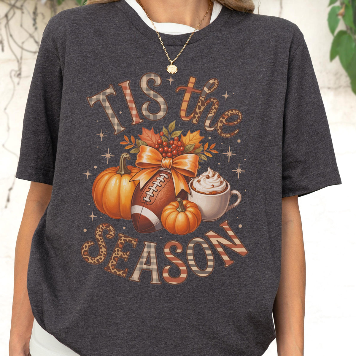 May include: Dark gray t-shirt with a fall-themed graphic. The design includes pumpkins, a football, autumn leaves, and a cup of whipped cream. The text "TIS the SEASON" is displayed in a decorative font.
