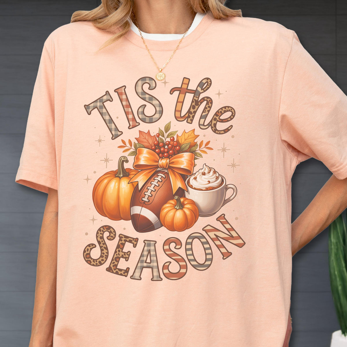 May include: Peach-colored t-shirt with a fall-themed graphic. The design includes pumpkins, a football, a coffee cup, and the text "Tis the Season". The text and graphic elements have a mix of patterns and colors.