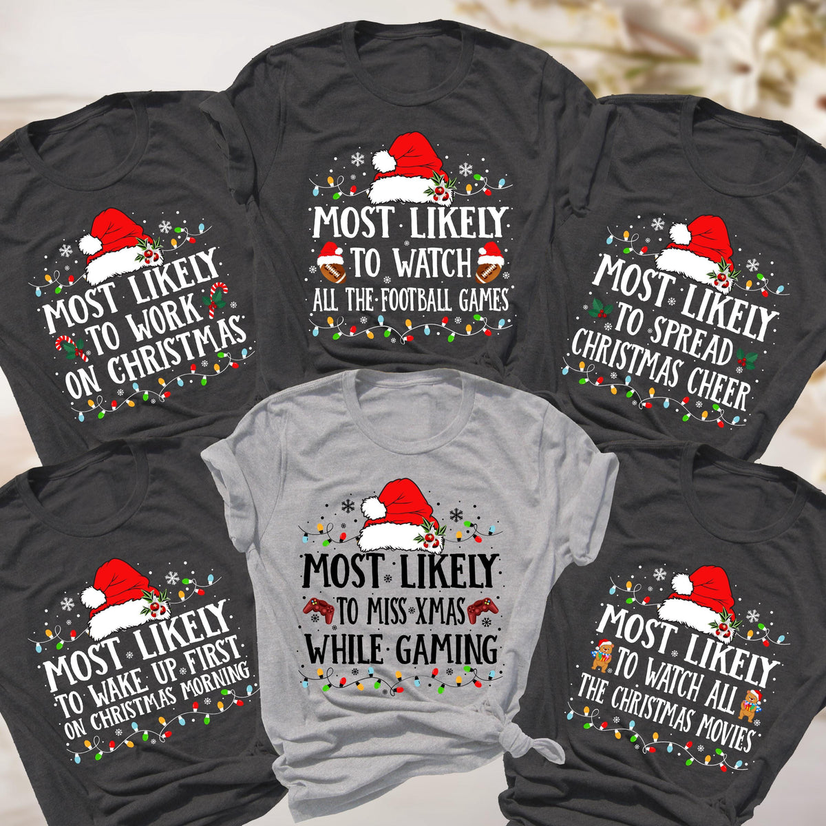 May include: A collection of gray and light gray t-shirts with Christmas-themed text and graphics. Each shirt features a Santa hat, string lights, and text like "Most Likely to Work on Christmas" or "Most Likely to Watch All the Christmas Movies."