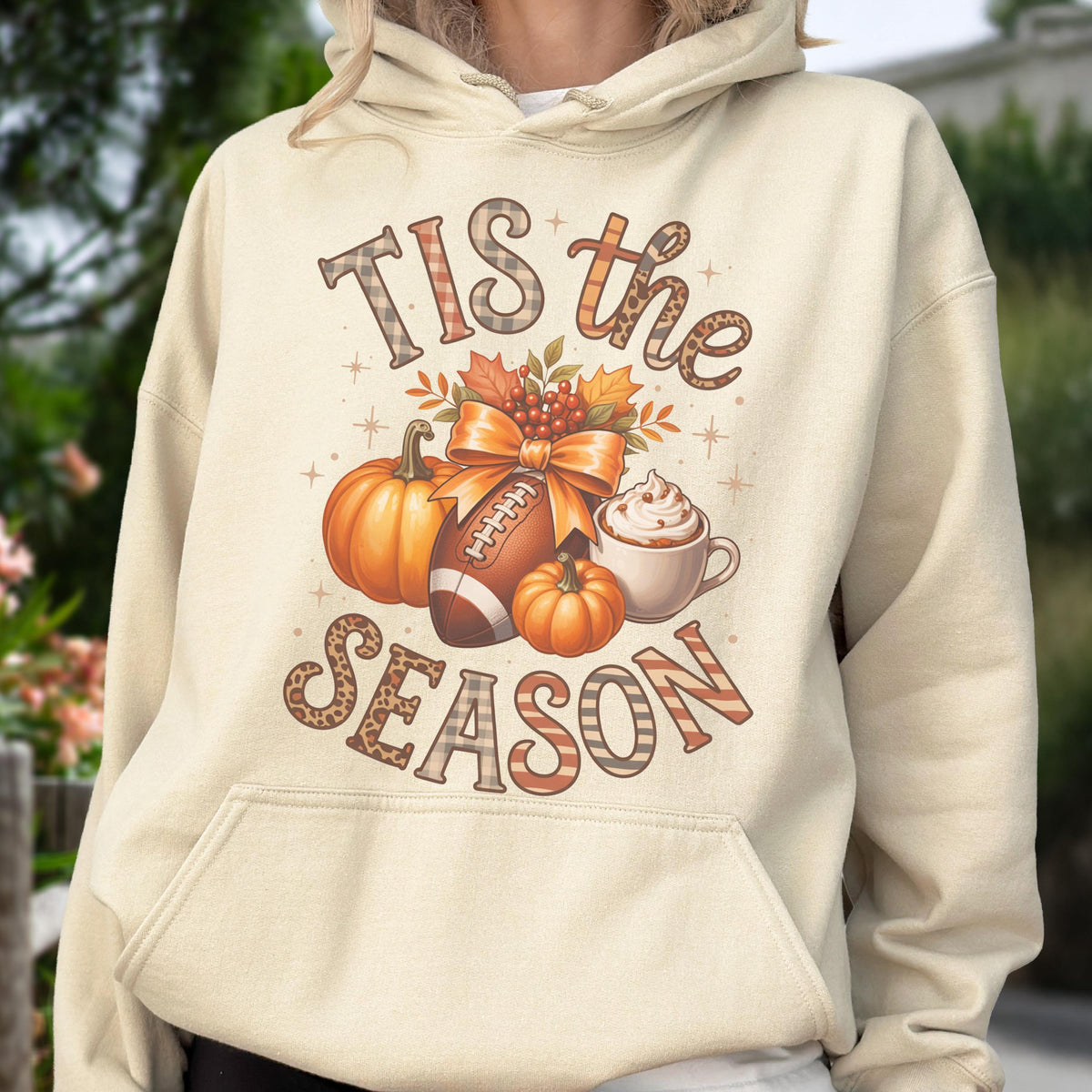 May include: Cream-colored hoodie with a fall-themed graphic. The design includes pumpkins, a football, a bow, and a coffee cup. The text "Tis the Season" is displayed in a mix of patterns and fonts, perfect for autumn.