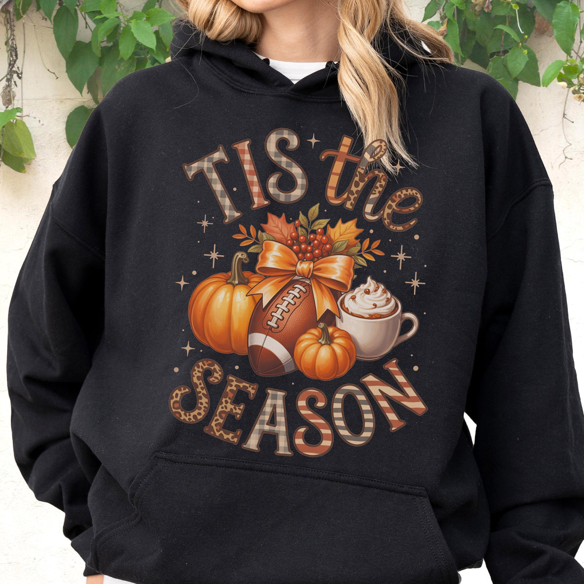 May include: Black hoodie with a fall-themed graphic. The design includes pumpkins, a football, and a cup of whipped cream. The text "TIS the SEASON" is displayed in a mix of patterns and fonts.