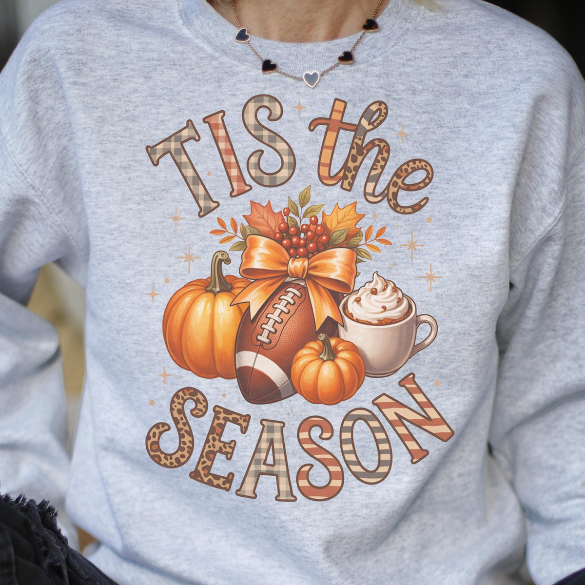 May include: A light gray sweatshirt with a fall-themed graphic. The graphic includes pumpkins, a football, a bow, and a cup of whipped cream. The text reads "Tis the Season" in a mix of plaid and leopard print.