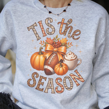 May include: A light gray sweatshirt with a fall-themed graphic. The graphic includes pumpkins, a football, a bow, and a cup of whipped cream. The text reads "Tis the Season" in a mix of plaid and leopard print.
