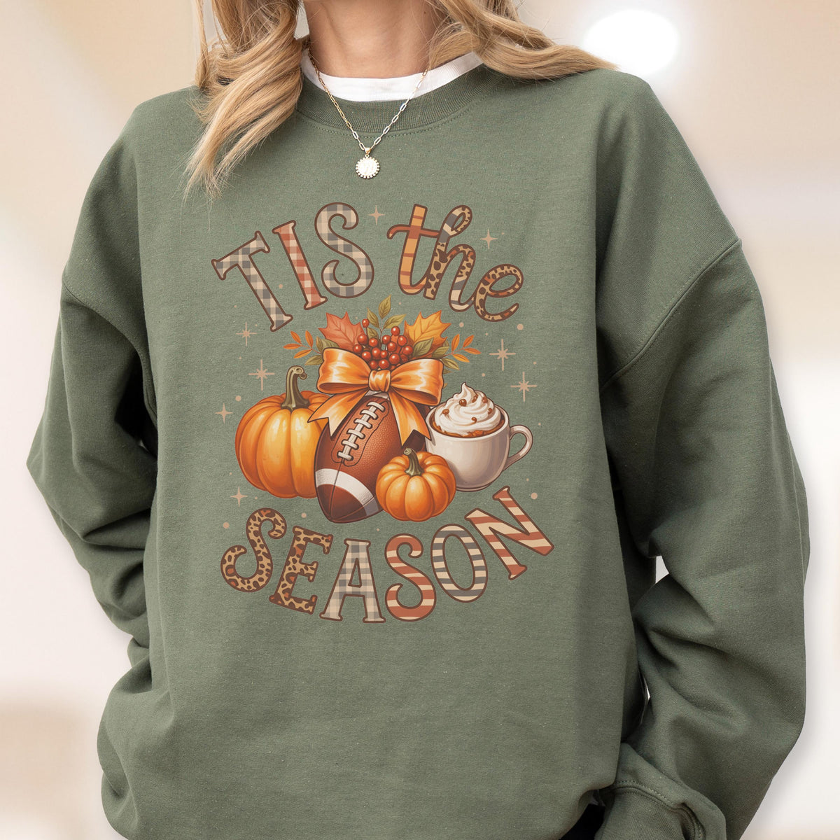 May include: Green sweatshirt with a fall-themed graphic. The design includes pumpkins, a football, a bow, and a cup of whipped cream. The text "Tis the Season" is displayed in a decorative font.