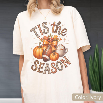 May include: Ivory-colored t-shirt with a fall-themed graphic. The design includes pumpkins, a football, and a coffee cup, with the text "Tis the Season" in decorative lettering. The shirt is a relaxed fit.