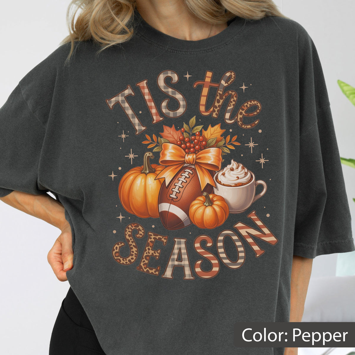 May include: Pepper-colored t-shirt with a fall-themed graphic. The design includes pumpkins, a football, and a coffee cup, with the text "TIS the SEASON" in various patterns and fonts. The shirt has a relaxed fit.