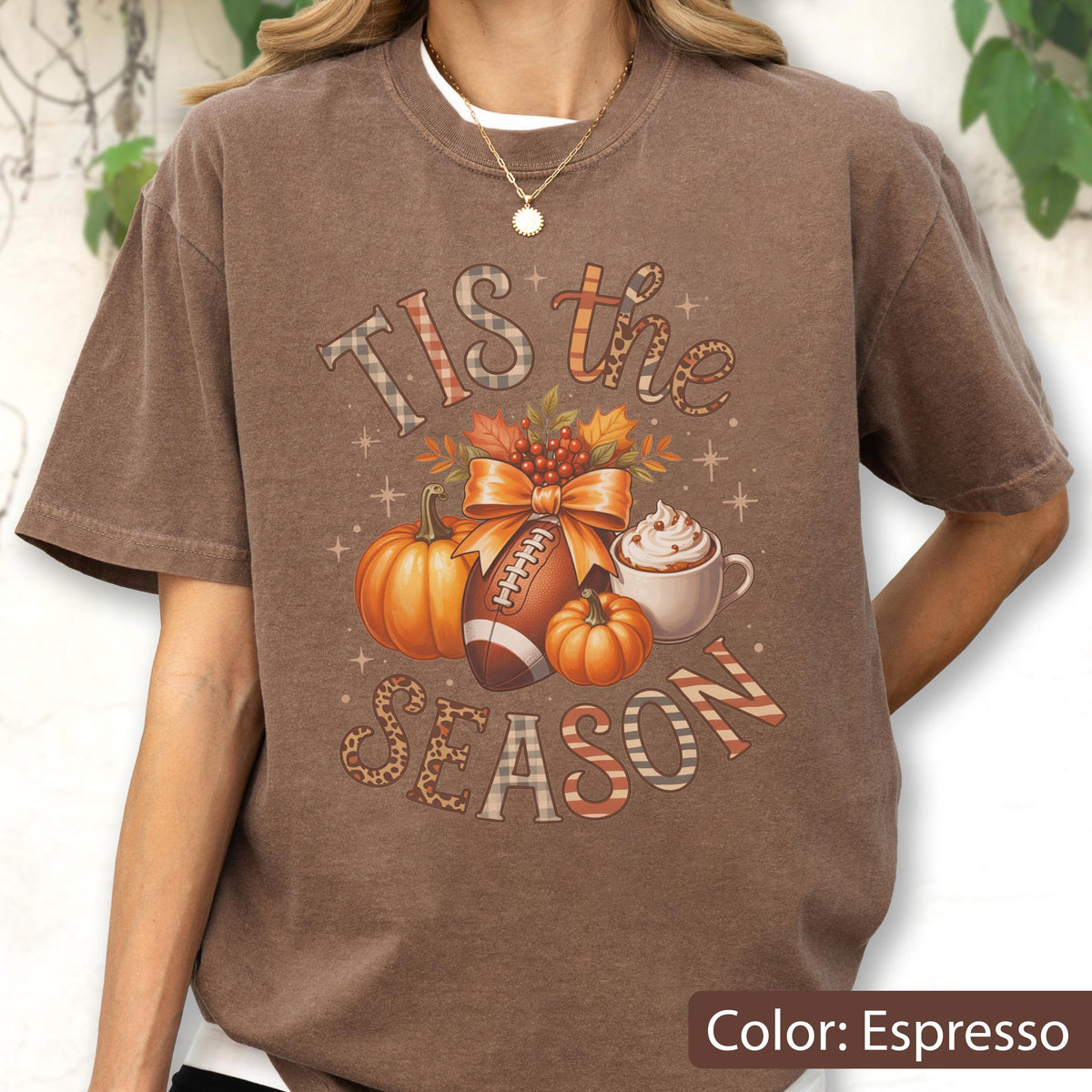 May include: Espresso-colored t-shirt with a fall-themed graphic. The design includes pumpkins, a football, and a cup of whipped cream. The text reads "Tis the Season" in a mix of fonts and patterns, perfect for autumn.