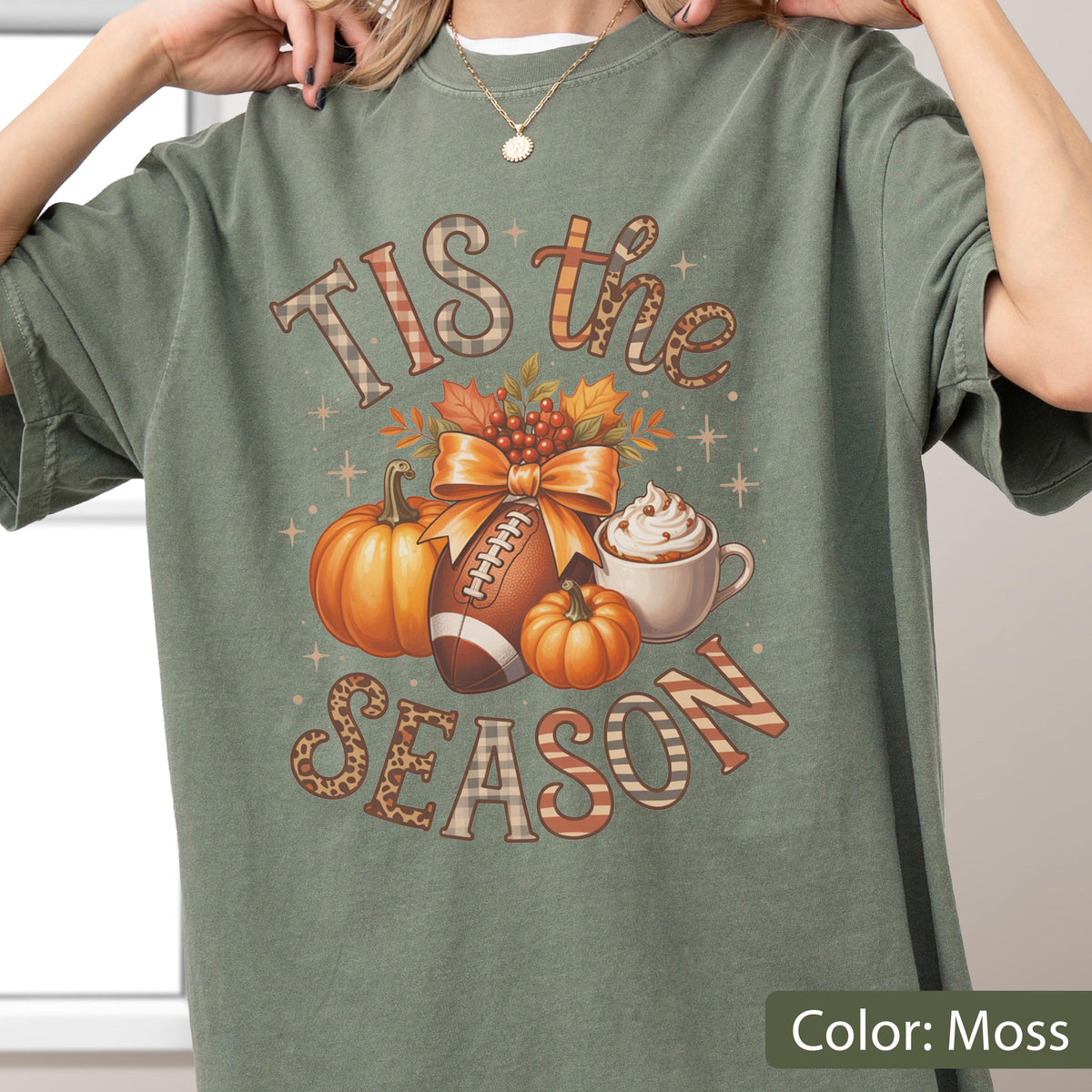 May include: Moss green t-shirt with a fall-themed graphic. The design includes pumpkins, a football, a bow, and the text "Tis the Season." A cup of whipped cream is also featured in the design.