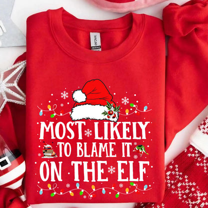 May include: A bright red crewneck sweatshirt featuring a graphic of a Santa hat with holly berries. The text reads MOST LIKELY TO BLAME IT ON THE ELF in white capital letters, surrounded by colorful Christmas light bulbs and white snowflakes.