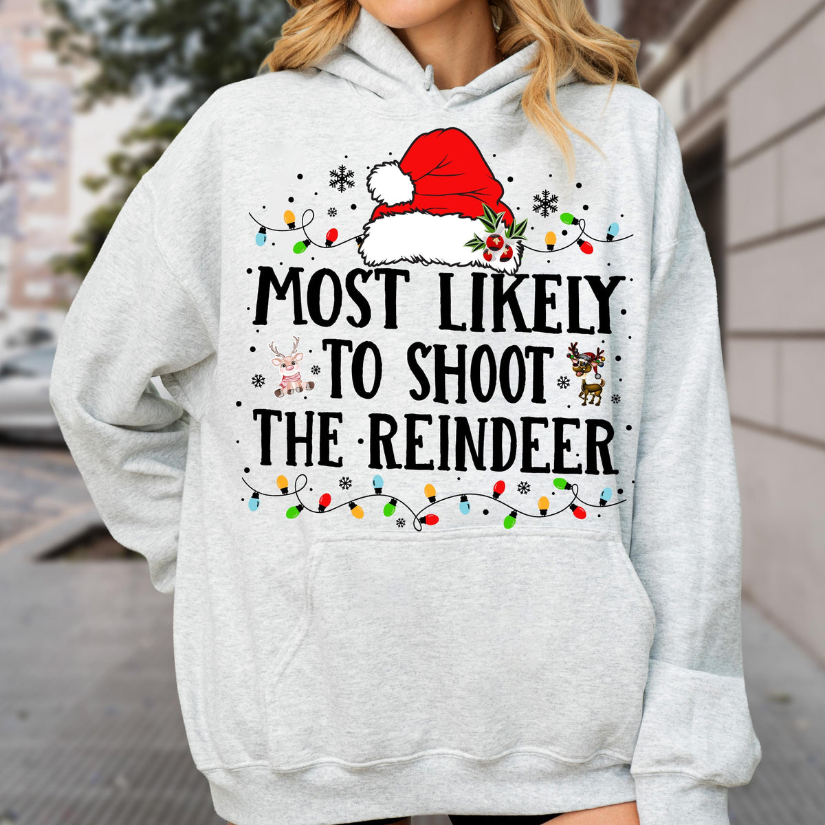 May include: A light gray hooded sweatshirt featuring a red Santa hat graphic at the top, colorful Christmas light strings, and the black text MOST LIKELY TO SHOOT THE REINDEER. Small reindeer illustrations are positioned near the text.