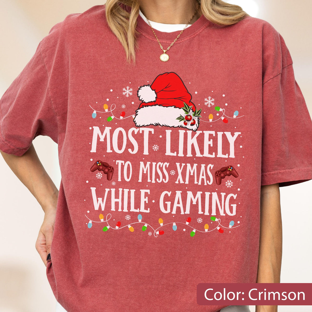 May include: A crimson short-sleeve t-shirt featuring a white graphic of a Santa hat, two red game controllers, and colorful Christmas lights. The text reads: MOST LIKELY TO MISS XMAS WHILE GAMING. Small white snowflakes are scattered throughout the design.