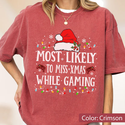 May include: Crimson-colored t-shirt with the text "MOST LIKELY TO MISS XMAS WHILE GAMING." The design includes a Santa hat, snowflakes, game controllers, and Christmas lights. A festive holiday-themed tee.