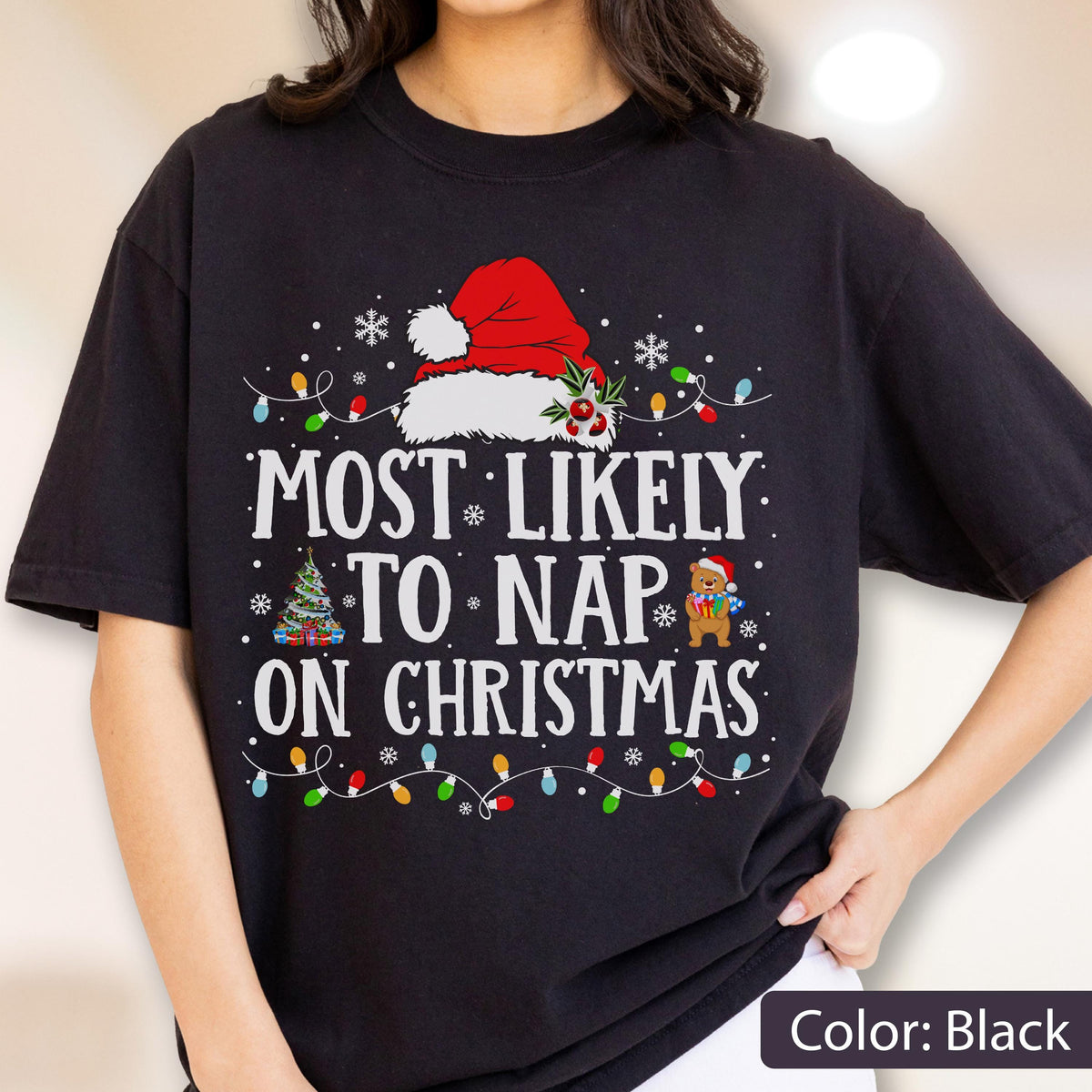 May include: A black short-sleeved t-shirt featuring a graphic with the text "MOST LIKELY TO NAP ON CHRISTMAS" in white. The design includes a red Santa hat, a decorated Christmas tree, a small bear holding a gift, and colorful string lights.
