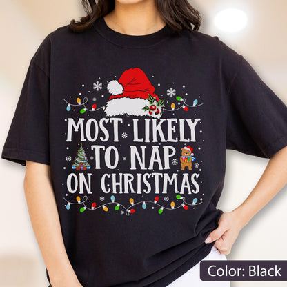 May include: A black short-sleeved t-shirt featuring a graphic with the text "MOST LIKELY TO NAP ON CHRISTMAS" in white. The design includes a red Santa hat, a decorated Christmas tree, a small bear holding a gift, and colorful string lights.