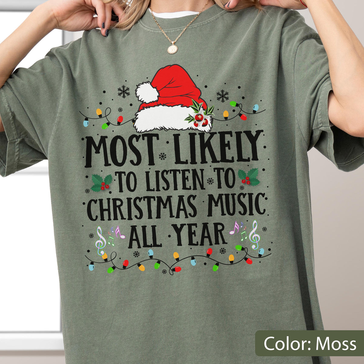 May include: A moss green t-shirt featuring a graphic of a red Santa hat with holly, surrounded by colorful string lights, snowflakes, and musical notes. The text reads: MOST LIKELY TO LISTEN TO CHRISTMAS MUSIC ALL YEAR in black capital letters.
