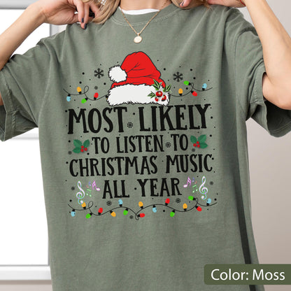 May include: A moss green t-shirt featuring a graphic of a red Santa hat with holly, surrounded by colorful string lights, snowflakes, and musical notes. The text reads: MOST LIKELY TO LISTEN TO CHRISTMAS MUSIC ALL YEAR in black capital letters.