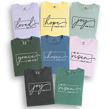 May include: A collection of t-shirts in various colors, including lavender, yellow, and green. Each shirt features a different word in a handwritten font, such as "loved," "hope," and "joy," inside a rectangular frame. The shirts also include Bible verses.