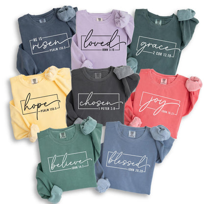 May include: A collection of eight crewneck sweatshirts in various colors, including blue, purple, yellow, and coral. Each sweatshirt features a different word in a white script font, such as "loved" and "joy", with a corresponding Bible verse.