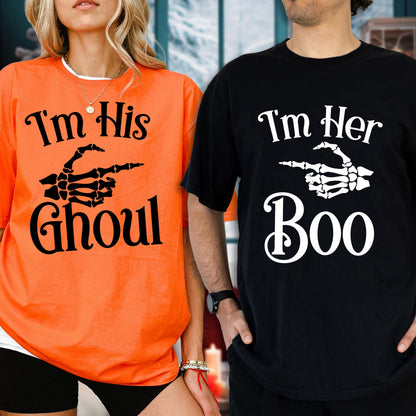 May include: An orange t-shirt with black text reading I'm His Ghoul and a black skeleton hand graphic, and a black t-shirt with white text reading I'm Her Boo and a white skeleton hand graphic, both featuring short sleeves and crew necklines.