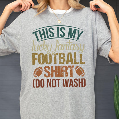 May include: A heather gray short-sleeve t-shirt featuring the text "THIS IS MY lucky fantasy FOOTBALL SHIRT (DO NOT WASH)" in dark green, tan, and brown, accompanied by two brown football illustrations on the front of the garment.