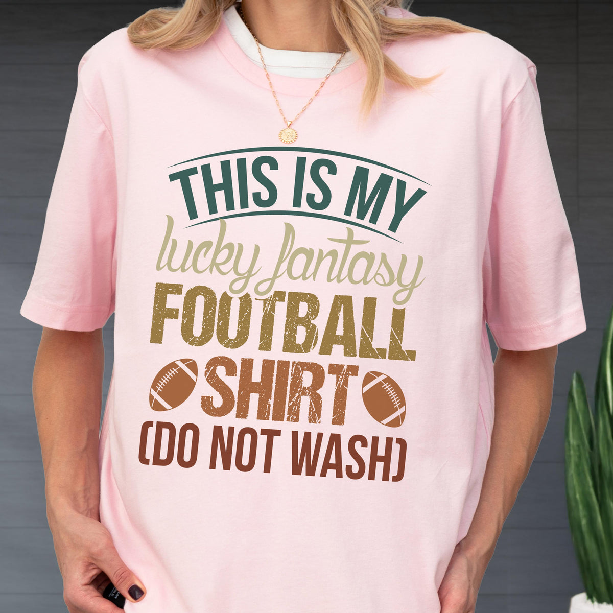 May include: A light pink short-sleeve t-shirt featuring the dark green and brown text THIS IS MY lucky fantasy FOOTBALL SHIRT (DO NOT WASH) with two brown football graphics. The design is centered on the chest of the crew neck garment.