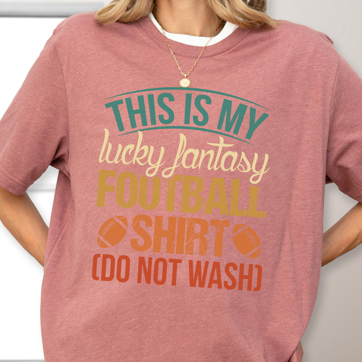 May include: A dusty rose short-sleeved t-shirt featuring the text "THIS IS MY lucky fantasy FOOTBALL SHIRT (DO NOT WASH)" in teal, cream, yellow, and orange, with two small football graphics flanking the word "SHIRT" on the front.