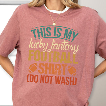 May include: A dusty rose short-sleeved t-shirt featuring the text "THIS IS MY lucky fantasy FOOTBALL SHIRT (DO NOT WASH)" in teal, cream, yellow, and orange, with two small football graphics flanking the word "SHIRT" on the front.