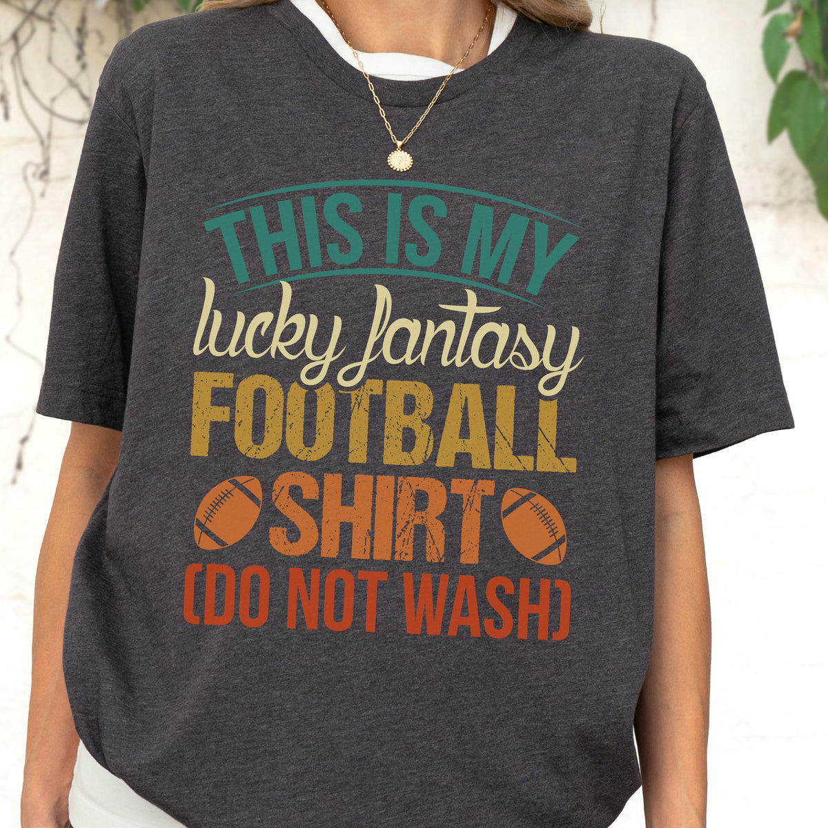May include: A dark charcoal gray short-sleeve t-shirt featuring the text "THIS IS MY lucky fantasy FOOTBALL SHIRT (DO NOT WASH)" in teal, cream, yellow, and orange, accompanied by two brown football illustrations on the front of the garment.