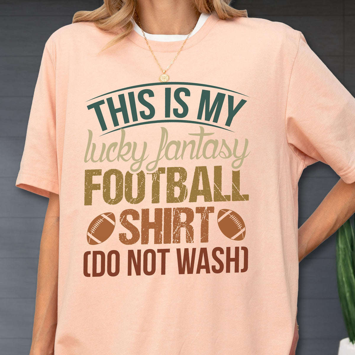 May include: A peach-colored t-shirt featuring the dark green and brown text THIS IS MY lucky fantasy FOOTBALL SHIRT (DO NOT WASH) with two brown football illustrations on the front, worn with a thin gold chain necklace.