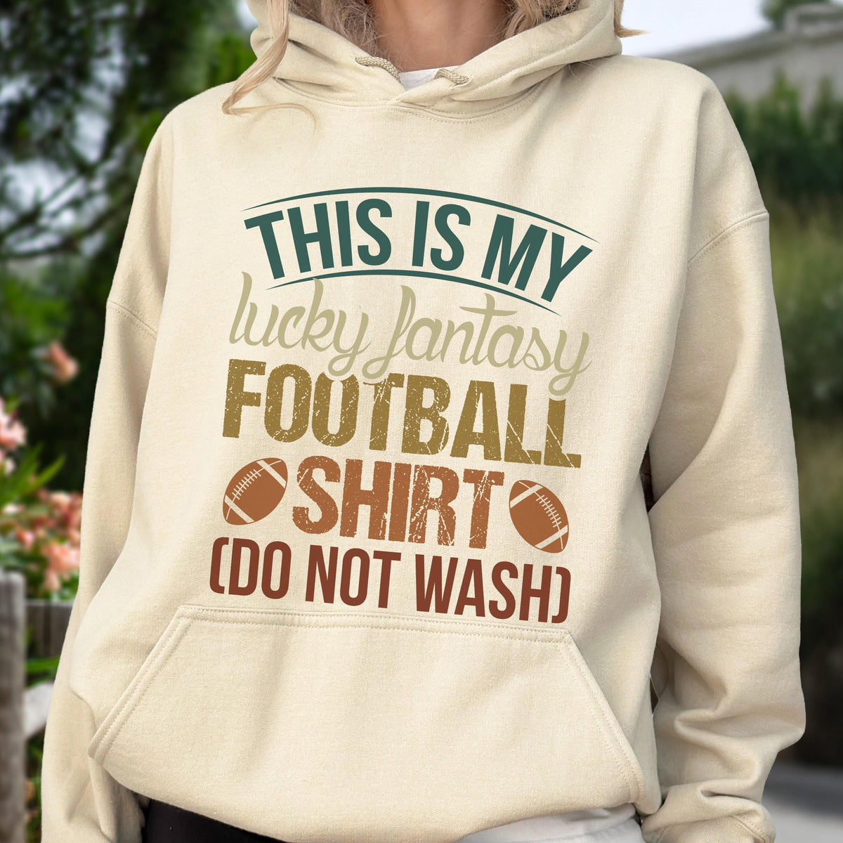May include: A beige hoodie with the text "THIS IS MY lucky fantasy FOOTBALL SHIRT (DO NOT WASH)" in green, brown, and beige. The design includes two football graphics. The hoodie has a front pocket and a drawstring hood.