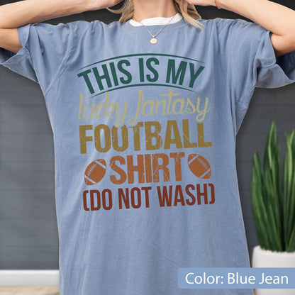 May include: A blue jean colored t-shirt featuring the text THIS IS MY lucky fantasy FOOTBALL SHIRT (DO NOT WASH) in dark green, yellow, and brown lettering, accompanied by two brown football graphics on the front of the garment.