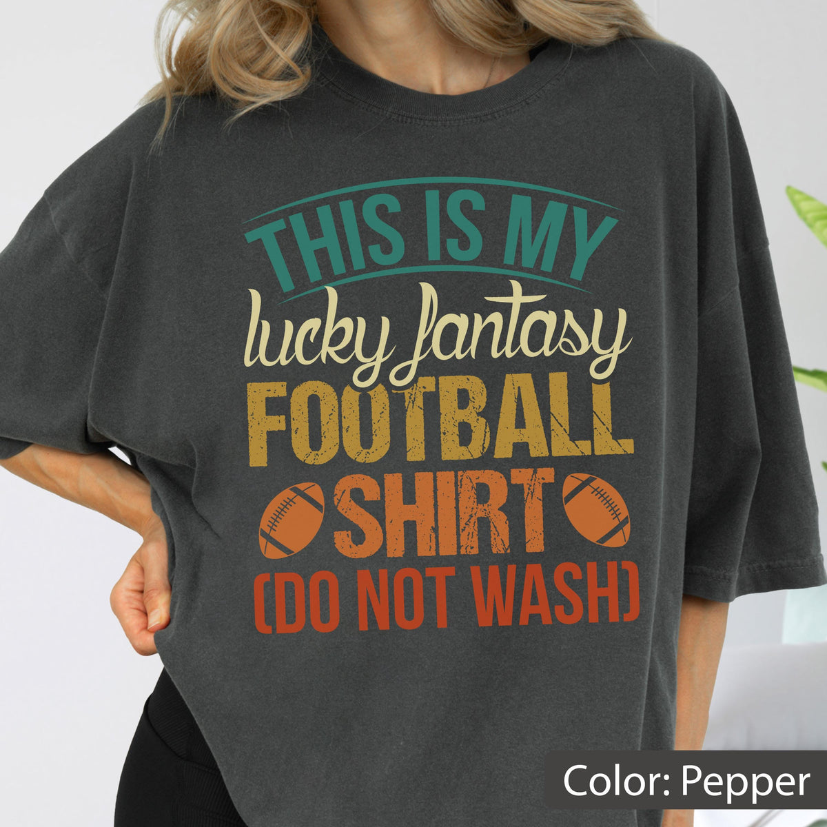 May include: A charcoal gray t-shirt featuring a graphic print with the text "THIS IS MY lucky fantasy FOOTBALL SHIRT (DO NOT WASH)" in teal, cream, yellow, and orange, accompanied by two brown football illustrations on the front of the garment.
