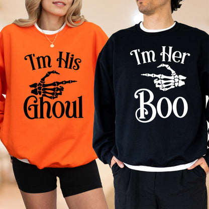 May include: Two sweatshirts, one orange and one black, with Halloween-themed text and graphics. The orange sweatshirt says "I'm His Ghoul" with a skeleton hand graphic. The black sweatshirt says "I'm Her Boo" with a skeleton hand graphic.