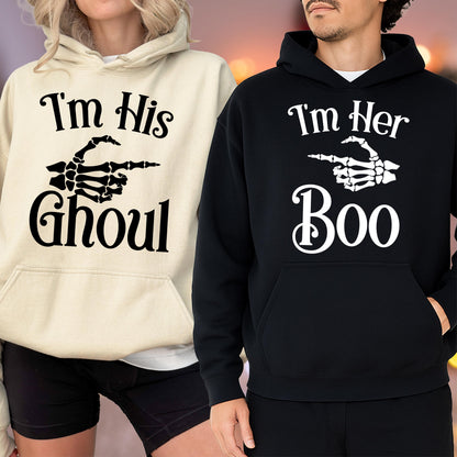May include: Two hooded sweatshirts, one cream and one black, with Halloween-themed graphics. The cream sweatshirt says "I'm His Ghoul" with a skeleton hand pointing. The black sweatshirt says "I'm Her Boo" with a skeleton hand pointing.
