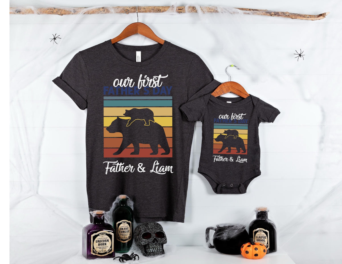 May include: Two t-shirts, one adult size and one baby size, both dark gray with a retro-style graphic of a bear family with the text 'our first father's day' and 'Father & Liam'.
