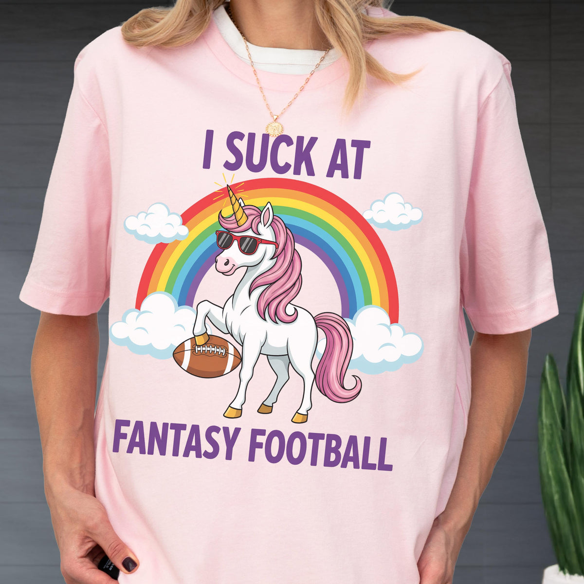 May include: A light pink t-shirt featuring a graphic of a white unicorn with a pink mane, wearing sunglasses, holding a brown football under one hoof. Above the unicorn is a rainbow and clouds, with the text I SUCK AT FANTASY FOOTBALL in purple.