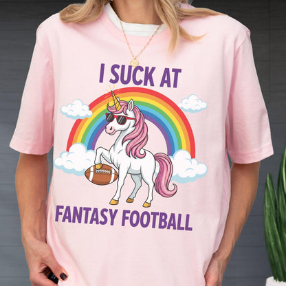 May include: A light pink t-shirt featuring a graphic of a white unicorn with a pink mane, wearing sunglasses, holding a brown football under one hoof. Above the unicorn is a rainbow and clouds, with the text I SUCK AT FANTASY FOOTBALL in purple.