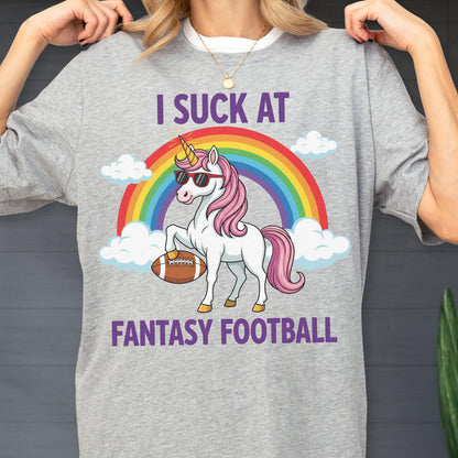 May include: A heather gray t-shirt featuring a graphic of a white unicorn with a pink mane and tail, wearing red sunglasses, holding a brown football. Behind the unicorn is a colorful rainbow and white clouds. The text reads I SUCK AT FANTASY FOOTBALL.