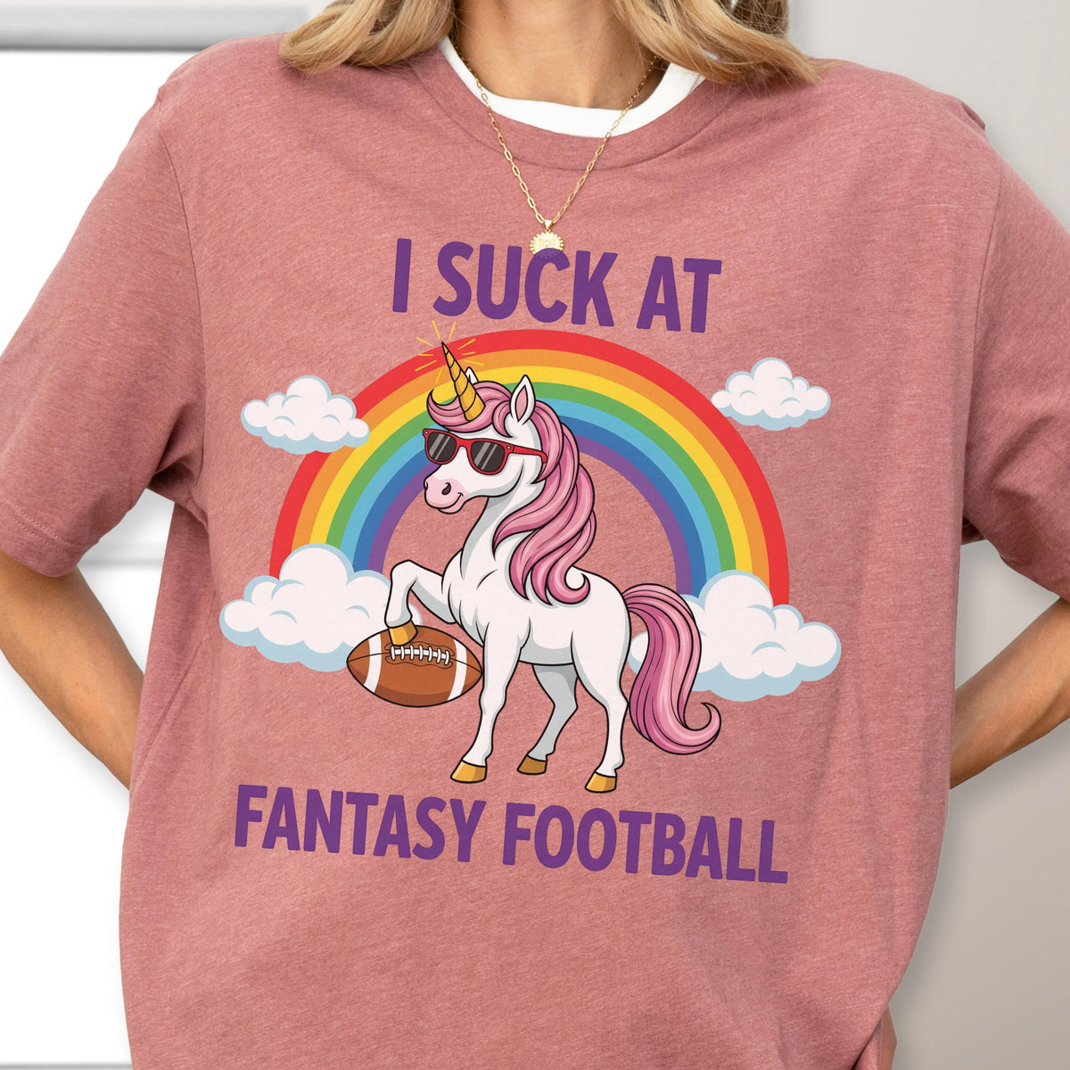 May include: A dusty rose t-shirt featuring a graphic of a white unicorn with a pink mane, wearing sunglasses and holding a brown football. Behind the unicorn is a colorful rainbow and white clouds. The text reads I SUCK AT FANTASY FOOTBALL.