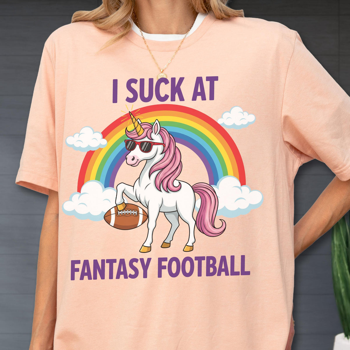 May include: A peach-colored t-shirt featuring a graphic of a white unicorn with a pink mane and sunglasses, holding a brown football. Above and below the unicorn, the text reads I SUCK AT FANTASY FOOTBALL, set against a colorful rainbow and clouds.