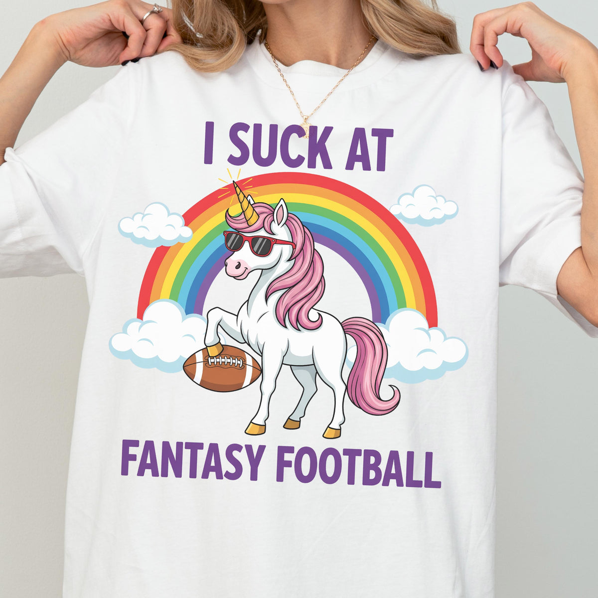 May include: A white t-shirt featuring a graphic of a white unicorn with a pink mane, wearing black sunglasses, holding a brown football, and standing in front of a colorful rainbow and white clouds with the text I SUCK AT FANTASY FOOTBALL.