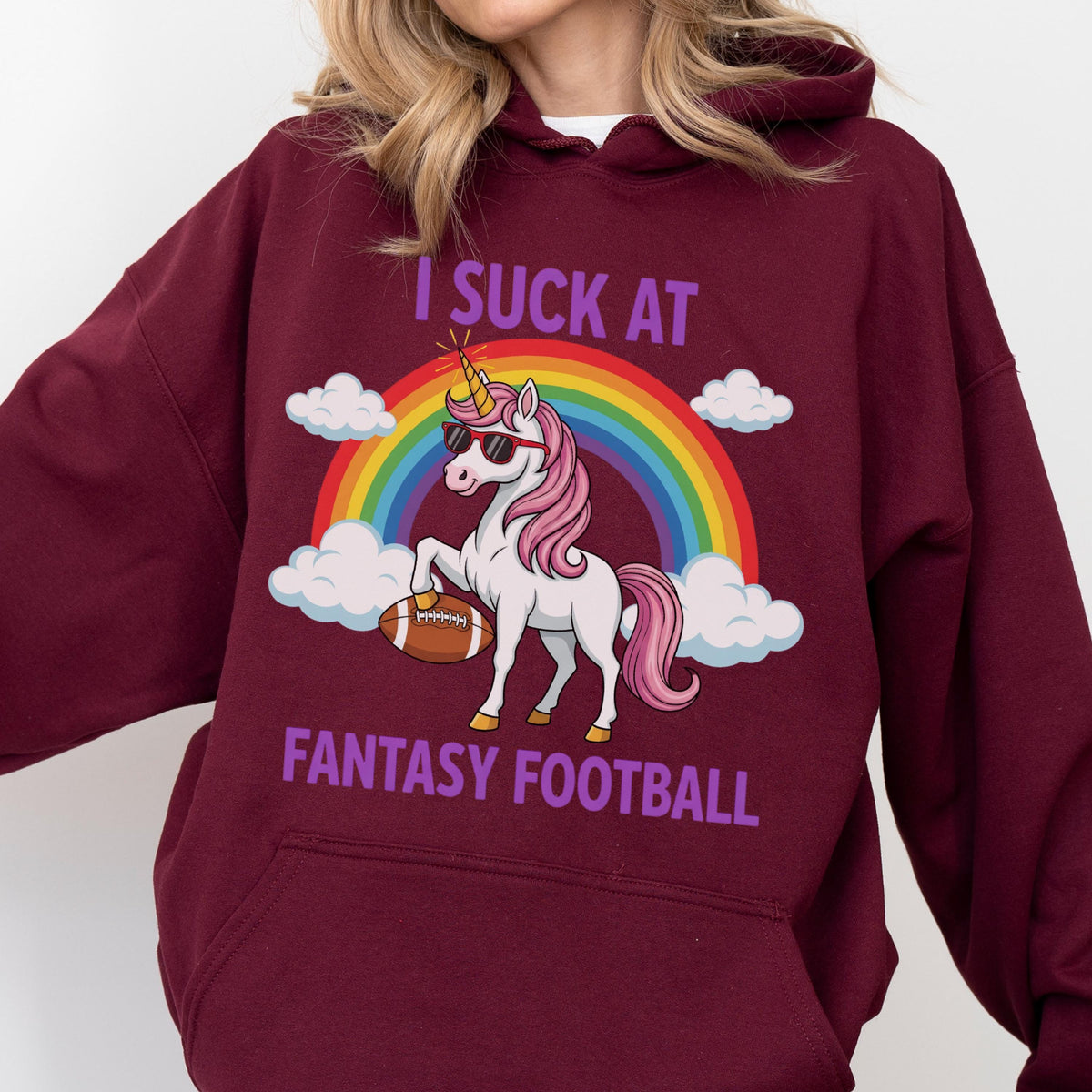 May include: A maroon hooded sweatshirt featuring a graphic of a white unicorn with a pink mane and tail, wearing sunglasses, holding a brown football under a rainbow and clouds. The text reads I SUCK AT FANTASY FOOTBALL in purple capital letters.