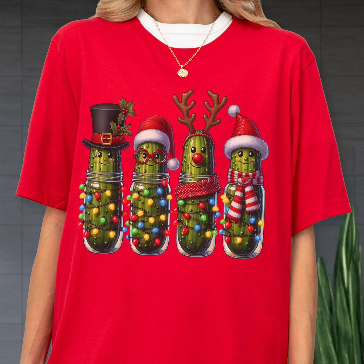 May include: Red t-shirt featuring a festive design of four anthropomorphic pickles in mason jars, each adorned with holiday accessories like a top hat, Santa hat, reindeer antlers, and scarves. The pickles are decorated with colorful Christmas lights.