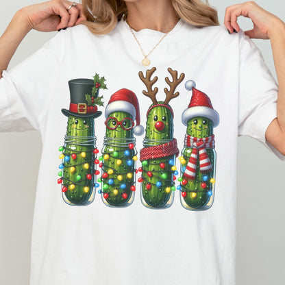 May include: White t-shirt featuring a festive graphic of four anthropomorphic pickles in glass jars, each adorned with holiday accessories like a top hat, Santa hat, reindeer antlers, and scarves. The pickles are decorated with colorful string lights.