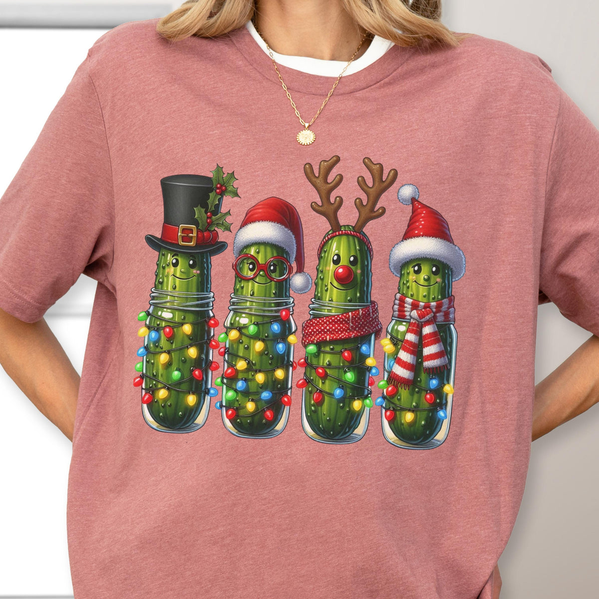 May include: A pink t-shirt featuring a whimsical illustration of four pickles in glass jars, each adorned with festive holiday accessories. The pickles are decorated with Christmas lights, hats, and scarves, creating a cheerful holiday theme.