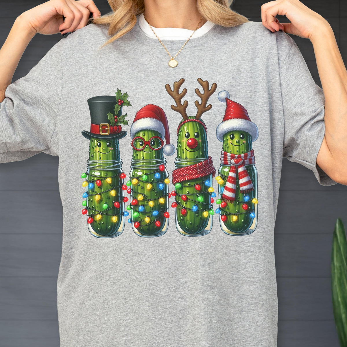 May include: A light gray t-shirt featuring a festive design of four anthropomorphic pickles in glass jars. Each pickle is decorated with holiday accessories like a top hat, Santa hat, reindeer antlers, and a scarf, all adorned with colorful string lights.