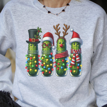 May include: A light gray crewneck sweatshirt featuring a graphic of four green pickles in glass jars. The pickles are decorated with Christmas themes including a top hat, Santa hats, reindeer antlers, a red nose, and colorful string lights.